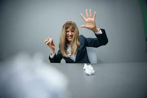 Funny millennial blonde businesswoman worker in blue jacket throws papers into the camera sitting at workplace in gray modern office. Tired of work Concept. Copy space, wide angle photo