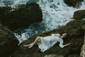 attractive woman with long hair in long white dress wet hair lying on a rocky cliff view from above photo