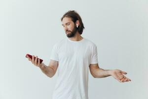 un hombre con un hipster barba en un blanco camiseta con un teléfono y inalámbrico auriculares sonriente escuchando a música y un audio libro en línea en contra un blanco pared foto