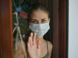 woman with a sad look in a medical mask looks out the window photo