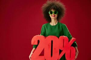 Positive young woman green dress afro hairstyle dark glasses twenty percent in hands red background unaltered photo