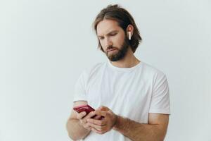 un hombre con un barba blogger en un blanco camiseta con un teléfono y inalámbrico auriculares mira dentro el teléfono y se extiende su manos a el lado en un blanco antecedentes aislado Copiar espacio foto
