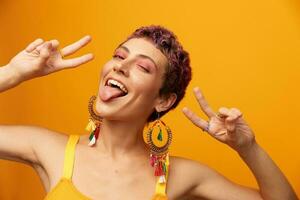 retrato de un joven mujer con un corto Corte de pelo y de colores pelo sonriente y demostración su lengua a el cámara en un naranja antecedentes con pendientes accesorios en el estudio foto