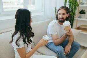 hombre y mujer sentado a hogar en el sofá en blanco elegante camisetas Bebiendo café fuera de cangrejo tazas desde un café tienda y teniendo divertido chateando sonrisas y la risa a hogar. masculino y hembra amistad foto