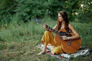 joven hippie mujer sentado al aire libre en un tartán y jugando su guitarra en el orilla del río dentro el puesta de sol foto