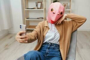 Happy funny blonde lady in pink fish mask warm sweater doing selfie video call sitting in armchair at modern home interior. Pause from work, take a break, social media in free time concept. Copy space photo