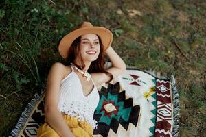 A woman in eco-friendly clothing in a hippie look is lying on a colored plaid smiling and looking at an autumn sunset in nature in the park. Lifestyle on a journey of harmony with the world photo