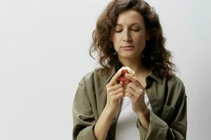 Happy cute curly beautiful woman in casual khaki green shirt eat apple with an appetite considers it posing isolated on over white background. Natural Eco-friendly products concept. Copy space photo