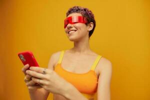 Woman blogger smiling in unusual red millennial glasses holding her phone and looking at the screen against an orange background in a stylish yellow top photo