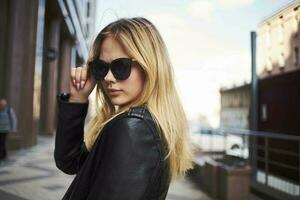 de moda mujer en un cuero chaqueta y Gafas de sol cerca el edificio foto