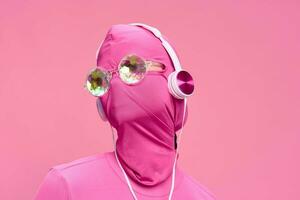 Portrait of a man wearing a pink full-face incognito mask with headphones and shiny round sunglasses on a pink background. Conceptual Art Photography Total Pink photo