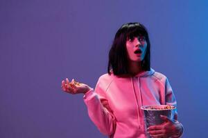 Shocked excited awesome brunet woman in pink hoodie with popcorn look aside open mouth posing isolated in blue violet color light studio background. Neon party Cyberpunk Cinema concept. Copy space photo