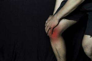 asiático hombre participación su rodilla. él siente dolor en su rodilla con negro antecedentes. articulación inflamación o cuidado de la salud concepto foto