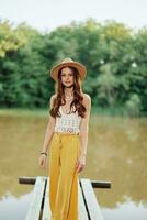 A young woman in a hippie look and eco-dress travels in nature by the lake wearing a hat and yellow pants in the fall sunset photo