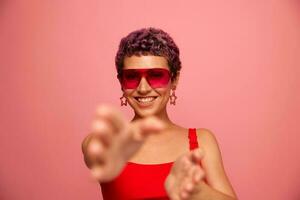 Fashion portrait of a woman with a short haircut in colored sunglasses with unusual accessories with earrings smiles on a pink bright background shows her hands to the camera photo