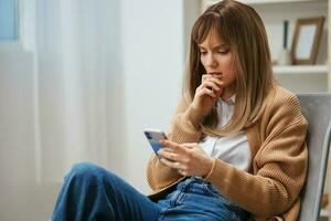 Confused unhappy young blonde lady in warm sweater read bad news use phone sitting in armchair at modern home interior. Pause from work, take a break, social media in free time concept. Copy space photo