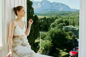 atractivo joven mujer admira el ver de el montañas en el abierto balcón de el hotel soleado día foto