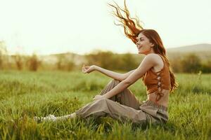 A woman sits on the green young fresh grass in the open air and laughs and smiles happily in the sunset light, her flying red hair fluttering in the wind photo