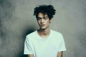 retrato de un hermoso chico con Rizado pelo en un gris antecedentes blanco camiseta de cerca modelo foto