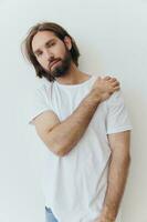 A man in a white T-shirt and blue jeans stands against a white wall and poses photo