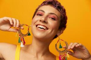 Portrait of a young woman with a short haircut and colored hair smiling and showing her tongue at the camera on an orange background with earrings accessories in the studio photo