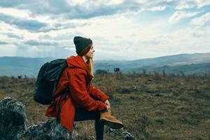 Traveler In a red jacket, hats with a backpack sits on a stone in the mountains in nature photo