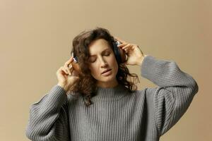 Pretty enjoyed curly beautiful female in gray casual sweater headphones listen favorite song posing isolated on over beige pastel background. Sound streaming platform concept. Copy space photo