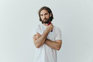 un hombre con un barba y largo pelo en un blanco camiseta y azul pantalones soportes en contra un blanco muro, propensión en contra eso y escuchando a música con inalámbrico blanco auriculares, curioso pensativamente foto