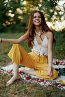 A young beautiful hippie woman sits on the ground in the fall in eco clothing in the sunset light and smiles sincerely for the camera photo