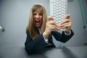 Crazy irritated angry millennial blonde businesswoman worker in blue jacket throws keyboard and screaming at camera in gray modern office. People Emotions Business Concept. Copy space, wide angle photo