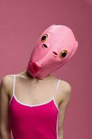 Woman wearing a pink fish head Halloween mask stands and looks at the camera against a pink background photo