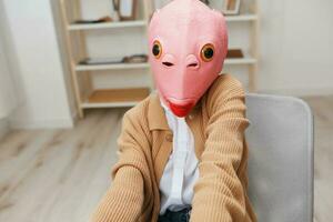 Cute funny blonde lady in pink fish mask warm sweater doing selfie use phone sitting in armchair at modern home interior. Pause from work, take a break, social media in free time concept. Wide angle photo