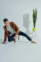 The guy is kneeling in a bright room near a flower and fashionable clothes style photo