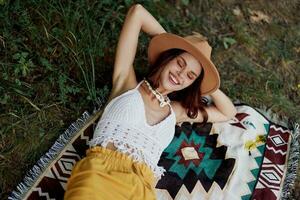 A woman in eco-friendly clothing in a hippie look is lying on a colored plaid smiling and looking at an autumn sunset in nature in the park. Lifestyle on a journey of harmony with the world photo