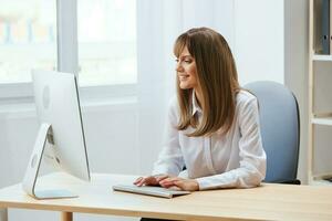 Cheerful happy adorable blonde businesswoman worker freelancer look at screen work on desktop computer online in light modern office. Happy employee typing quarterly report. Copy space, remote job photo