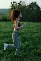 A woman with long red hair works out and runs on the green grass in the park in sweatpants and sneakers photo