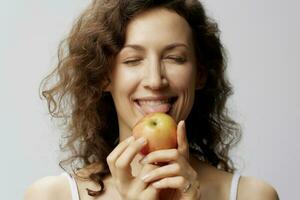 gracioso linda Rizado hermosa mujer en básico blanco camiseta disfrutar lame Fresco manzana sano comida posando aislado en terminado blanco antecedentes. natural Respetuoso del medio ambiente productos concepto. Copiar espacio foto