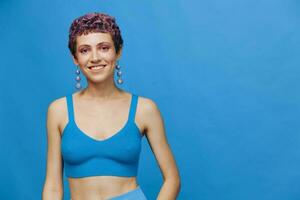 Young athletic fashion woman with colored hair and short haircut posing and dancing in blue sportswear smiling and looking at the camera on a blue monochrome background photo