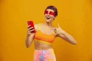 Woman blogger surprise in unusual millennial glasses taking selfies on her phone sporting brightly colored clothes against an orange studio background, free space photo