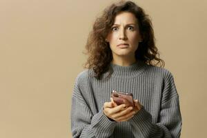 Confused unhappy curly lady in gray casual sweater read conflicting information in message posing isolated on over beige background. Social media, network, distance communication concept. Copy space photo