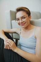 Teenage girl sitting on the floor at home smiling in home clothes and glasses with a short haircut, lifestyle without filters, free copy space photo