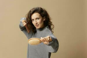 Funny awesome curly beautiful female in gray casual sweater hold hairbrush comb as weapon posing isolated on over beige pastel background. Problematic unruly damaged hair concept. Copy space photo