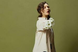 Sexy pretty curly beautiful female in linen casual shirt pulls chamomiles flowers at camera as gun sticks tongue out posing isolated on olive green background. Nature is beauty concept. Copy space photo