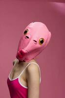 Woman wearing a pink fish head Halloween mask stands and looks at the camera against a pink background photo