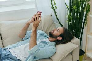 un hombre con un barba mentiras en el sofá durante el día a hogar y mira a su teléfono relajante en su día apagado, un hombre juego en el valores mercado en línea en su teléfono foto