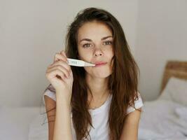 mujer participación un termómetro en su boca mientras sentado en el cama comprobación temperatura foto