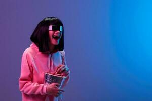 Laughing excited awesome brunet woman in pink hoodie trendy specular sunglasses eat popcorn look aside posing isolated in blue violet color light background. Neon party Cinema concept. Copy space photo