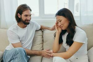 A man and a woman of different races sit on the couch in a room at home and talk about their problems to each other. A stress-free lifestyle of family quarrels with psychological support photo