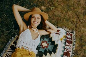 un mujer en Respetuoso del medio ambiente ropa en un hippie Mira es acostado en un de colores tartán sonriente y mirando a un otoño puesta de sol en naturaleza en el parque. estilo de vida en un viaje de armonía con el mundo foto