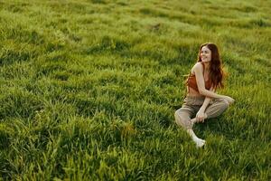 The sunset light of a summer evening illuminates a beautiful young redheaded woman sitting on the grass in green pants and an orange top and smiling at the sun photo